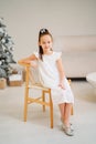 Cute little girl in white dress sitting on a chair by Christmas tree. Royalty Free Stock Photo