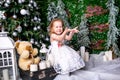 Cute little girl in a white dress sitting near a Christmas tree on a suitcase next to the candles and a teddy bear and throws snow Royalty Free Stock Photo