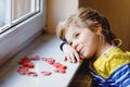 Cute little girl by window with lots of dough hearts as gift for Valentine& x27;s day, Mother& x27;s day or birthday