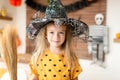 Cute little girl in witch costume with hat is holding a broom and standing in Halloween decorated living room, looking at camera. Royalty Free Stock Photo