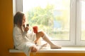 Cute little girl wrapped in warm plaid with cup of hot cocoa drink eating chocolate on windowsill at home