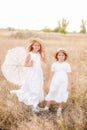 Cute little girls sisters with blond hair in a summer field at sunset in white dresses