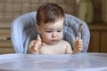 Cute little hungry baby boy holds spoon in his hand and looks at empty table waiting for food