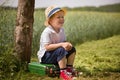 Cute little kid boy in shorts, white polo and straw hat sits on his green suitcase in a field near a tree waiting for a bus.