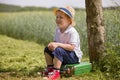 Cute little kid boy in shorts, white polo and straw hat sits on his green suitcase in a field and waiting for a bus