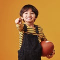 Cute little mixed race boy smiling and pointing while holding a football and posing against an orange copyspace Royalty Free Stock Photo