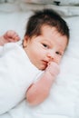 Cute little newborn baby with short dark hair in white bodysuit look sleepy at camera clenching fists. Royalty Free Stock Photo