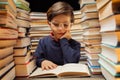 Cute little pupil boy in glasses reading interesting book in library between stacks of books literature. Education Royalty Free Stock Photo