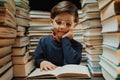 Cute little pupil boy in glasses reading book in library between stacks of books Royalty Free Stock Photo