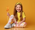 Cute little kid girl in yellow t-shirt and colorful leggings sits on floor holding big lollipop in hand and smiles Royalty Free Stock Photo