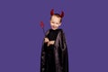 Cute little smiling boy on a purple background dressed in a black cloak with horns on his head and a trident in his hands looks at Royalty Free Stock Photo