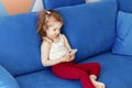 Cute little toddler girl wear casual clothes sitting on blue sofa and watching cartoon on smartphone Royalty Free Stock Photo