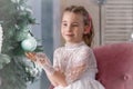 A cute little toddler girl in a white holiday dress sits near a festive New Year tree. Christmas Royalty Free Stock Photo