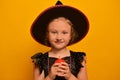 Little wizard girl in Halloween witch costume holding red burning candle and looking at camera on a yellow background.