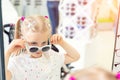Cute little young caucasian blond girl trying on and choosing sunglasses in front of mirror at optic eyewear store. Adorable Royalty Free Stock Photo