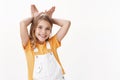 Cute lovely charming little girl, blond child in t-shirt, overalls, show bunny ears mimicking rabit, hold palms behind Royalty Free Stock Photo