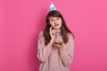 Cute lovely young woman dresses pink sweater, holding fake birthday cake with candles props, having birthday hat on her head, Royalty Free Stock Photo