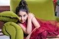 Cute lovlely middle eastern girl with dark red dress and collected hair posing and liying on green sofa at home interior.