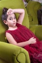 Cute lovlely middle eastern girl with dark red dress and collected hair posing and liying on green sofa at home interior.