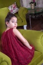 Cute lovlely middle eastern girl with dark red dress and collected hair posing and liying on green sofa at home interior.