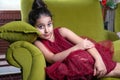 Cute lovlely middle eastern girl with dark red dress and collected hair posing and liying on green sofa at home interior.