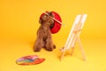 Cute Maltipoo in red beret holding brush near easel with canvas and palette on orange background. Dog artist Royalty Free Stock Photo