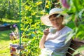 Cute mature female in a white hat is sitting in a beautiful summer garden with tea cup in her hands and drinking