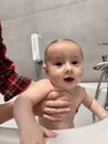 A cute mother is bathing a baby boy in clear water in a bathtub