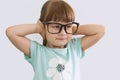 A cute naughty little girl with glasses covers her ears with her hands and smiles on a gray background. Children, the concept of e