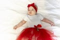a cute newborn baby girl is lying on white blanket dressed in a red skirt and with a red bow in a close-up view from Royalty Free Stock Photo