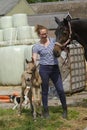 Cute newborn riding horse colt stands next to a woman in the grass. At the farmyard, yellow dun color. Dog behind the foal Royalty Free Stock Photo