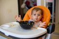 Cute one year old baby girl in an orange child seat in front of a plate of food throws up her hands and looks to the Royalty Free Stock Photo