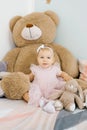 A cute one year old girl is sitting on the bed in the children`s room at home among soft funny toys in a pink dress