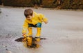 Cute playful little baby boy in bright yellow raincoat and rubber boots playing with rubber ducks in small puddle at rainy spring