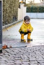 Cute playful little baby boy in bright yellow raincoat and rubber boots playing with rubber ducks in small puddle at rainy spring Royalty Free Stock Photo