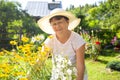 Cute positive senior woman in a white hat with a wide brim among flowers in summer garden