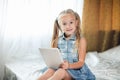 Cute pre teenager girl doing homework lying on bed at home. Young pretty girl wearing denim sundress studying online with tablet, Royalty Free Stock Photo