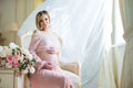 Cute pregnant woman sitting on chair near billowing curtains and hugs belly with love Royalty Free Stock Photo