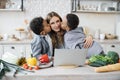 Cute preschool sons kissing their mother in light kitchen.
