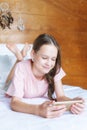 Cute preteen girl in pink t-shirt and shorts lying on bed with cell phone in boho style room against wooden wall and dreamcatchers