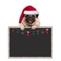 Cute pug puppy dog wearing santa hat with paws on blank blackboard sign with Christmas decoration Royalty Free Stock Photo