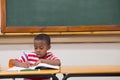 African American boy writing in notebook with pencil at classroom desk with textbook and chalkboard Royalty Free Stock Photo