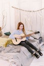 Cute red-haired woman learns to play the guitar, sitting on the bed, in a room in the style of boho