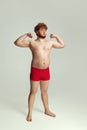 Cute red-headed man in red swimming shorts posing isolated on gray studio background. Concept of sport, humor and body