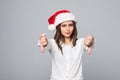 Cute sad Caucasian teenage girl wearing Santa hat and gloves showing thumbs down and making sad facial expression isolated on grey Royalty Free Stock Photo