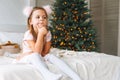 Cute sad funny little girl in pink dress with present gift box sitting on bed in room with Christmas tree. Portrait of unhappy Royalty Free Stock Photo