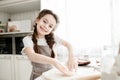 A cute school-age girl stands in the kitchen in an apron soiled in flour, smiles at the camera and rolls out the dough