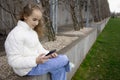 A cute schoolgirl girl is sitting on a bench in a spring park and watching videos or messages on her smartphone. Modern Royalty Free Stock Photo