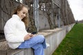 A cute schoolgirl girl is sitting on a bench in a spring park and watching videos or messages on her smartphone. Modern Royalty Free Stock Photo