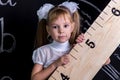 Cute schoolgirl standing before the chalkboard as a background holding the huge ruler diagonally. Enlarged view